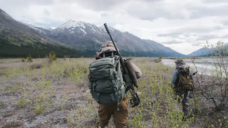 250610_SRME_AK_BEAR-104-1200x800-5b2df79jpg - PopularSuppressors Two hikers with large backpacks and rifles trek across a rocky valley toward snow-capped mountains in the distance.