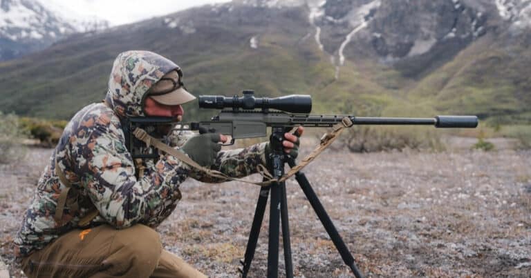Person in camouflage kneels in a rocky field, aiming a scoped rifle on a bipod with a tan sling, mountains in the background.