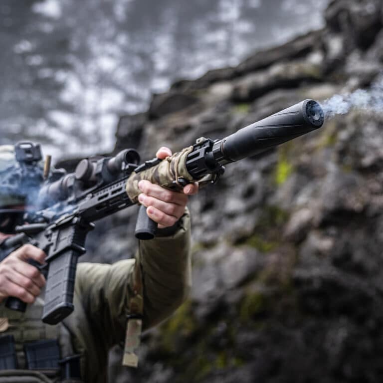 Person in camouflage firing a rifle with a suppressor outdoors on a rocky, mossy terrain.