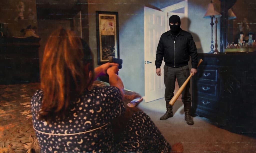 A masked man in a black jacket and balaclava stands in a doorway holding a wooden bat while a woman in starry pajama top points a gun at him from a couch in a dimly lit room.