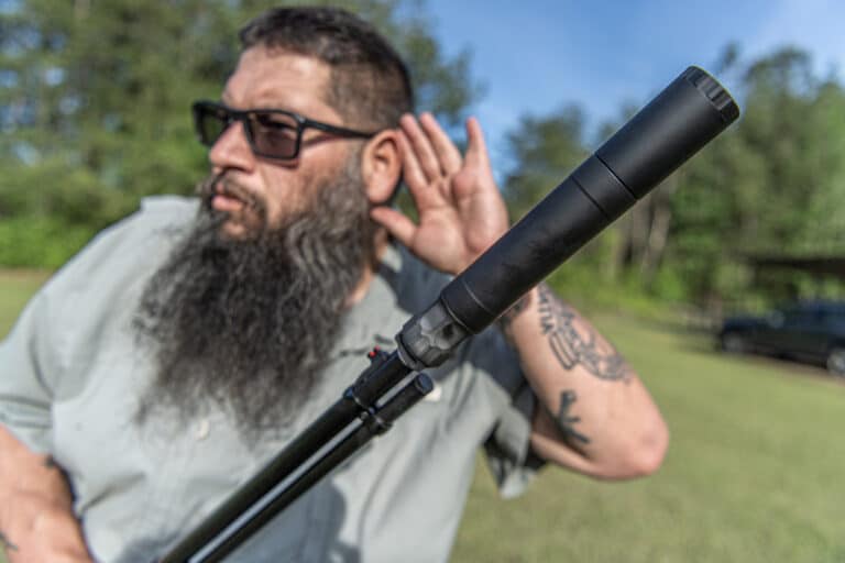 Bearded man wearing sunglasses outdoors, adjusting his ear while holding a black rifle with a suppressor.