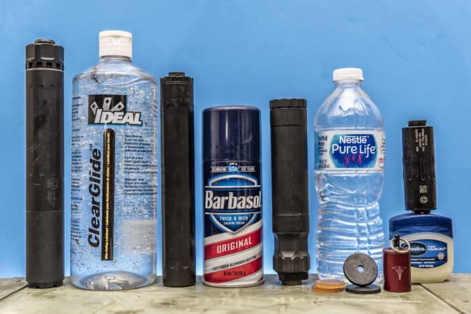 Row of household containers on a wooden table against a blue wall: a ClearGlide spray bottle, Barbasol Original shaving cream can, a bottle of Nestlé Pure Life water, and several black cylindrical items lined up in a row.