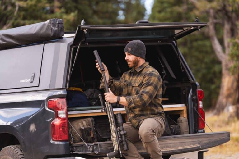 Man in a plaid shirt and beanie loading a rifle from the open bed of a pickup truck in a forested area.