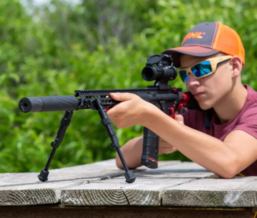 silencer central youth 3 - PopularSuppressors Young man wearing an orange cap and sunglasses aiming a scoped rifle with a bipod on a wooden table outdoors.