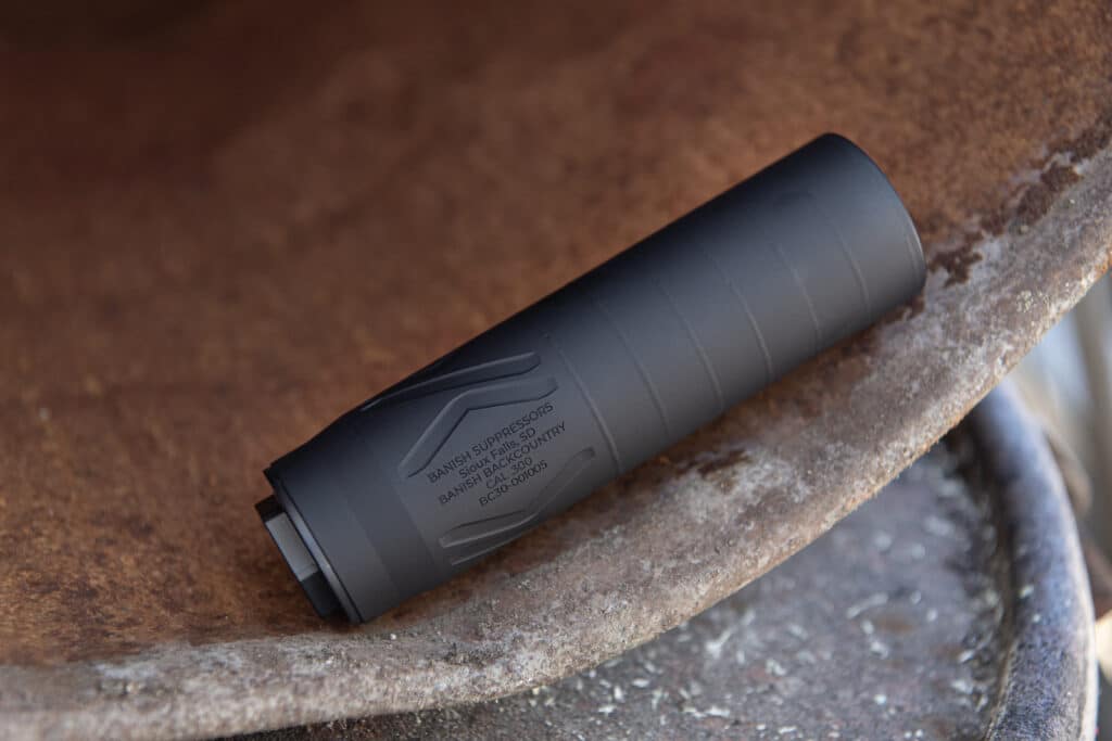 Close-up of a dark gray suppressor lying on a rusted metal surface, with unreadable stamped text along its body.