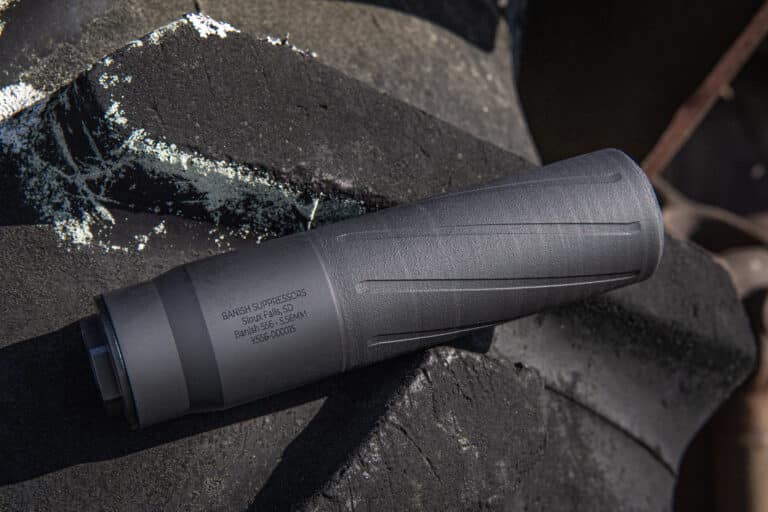 Close-up of a handgun suppressor attached to a barrel resting on charred rock. The device has stamped model text visible on its surface.