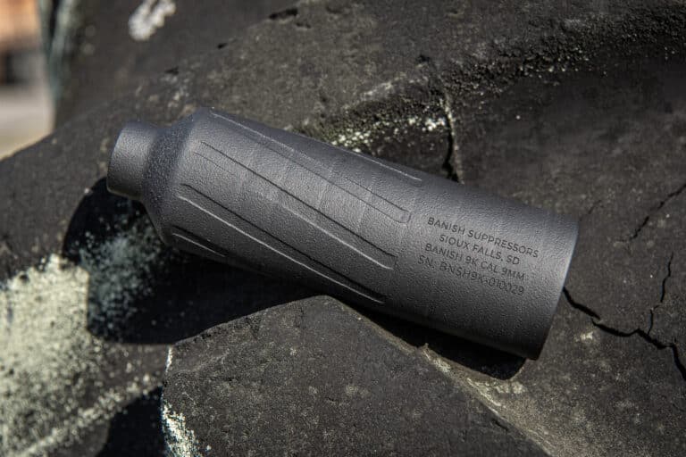 Close-up of a matte black firearm suppressor with engraved model and serial number on a rough, dark surface.