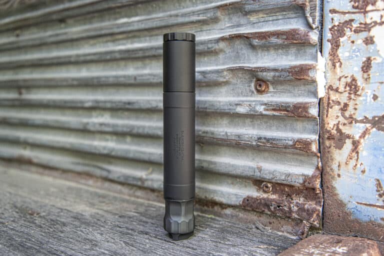 Black cylindrical firearm suppressor standing upright on a weathered wooden surface against a rusted, peeling metal wall.