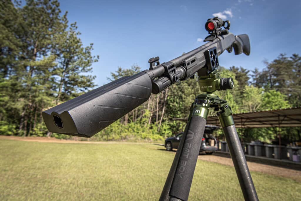 Close-up of a scoped rifle mounted on a bipod outdoors, aimed toward the sky with trees in the background.
