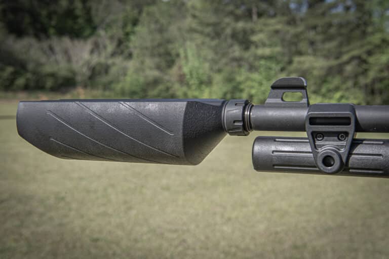 Close-up of a rifle stock and mounting rail outdoors with a blurred grassy background.