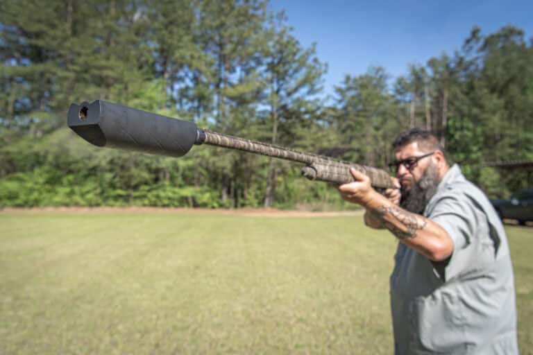 Man aiming a long-barreled shotgun outdoors, extending the gun toward the camera with a forested backdrop behind him.