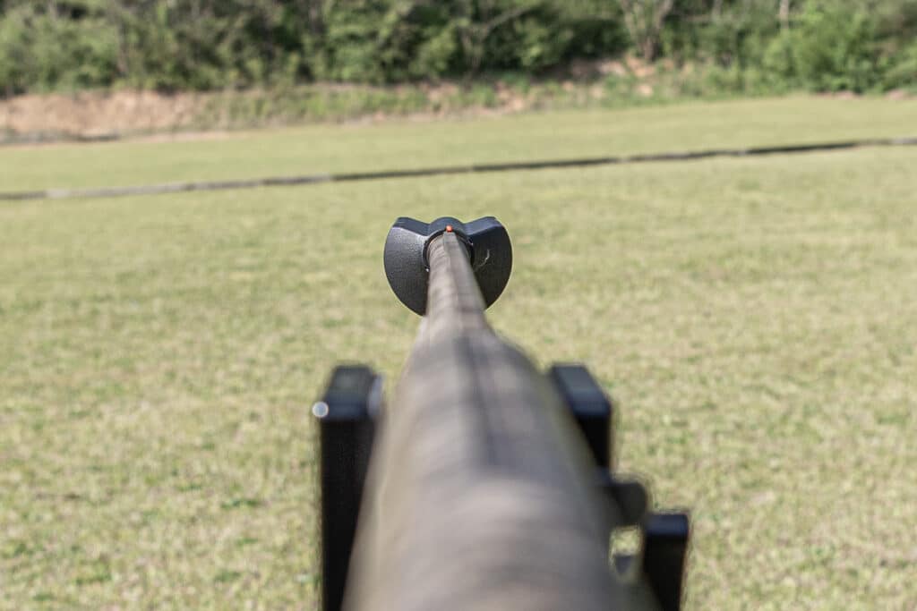 First-person view down the barrel of a rifle with a red-dot sight aimed at an open grassy field outdoors