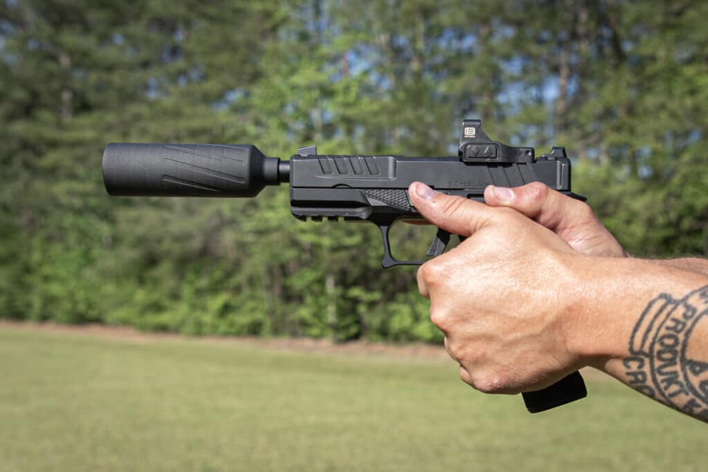 A pair of hands gripping a black semi-automatic pistol with a suppressor and red-dot sight, aimed Outdoors in a grassy area.