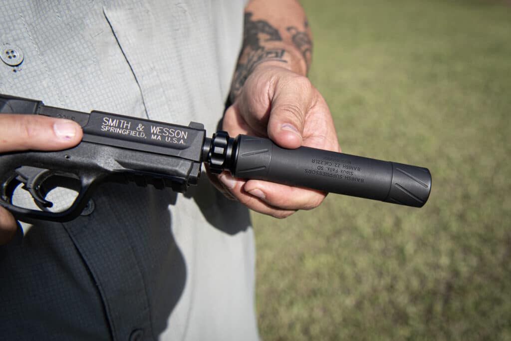 Close-up of a person holding a black Smith & Wesson rifle outdoors, with the brand visible on the receiver and a tattooed forearm nearby.