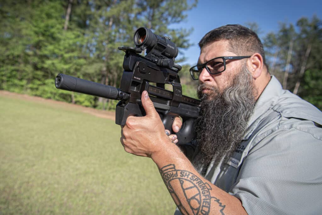 Man with a beard and glasses aiming a riflescope-equipped gun outdoors, close to the barrel.