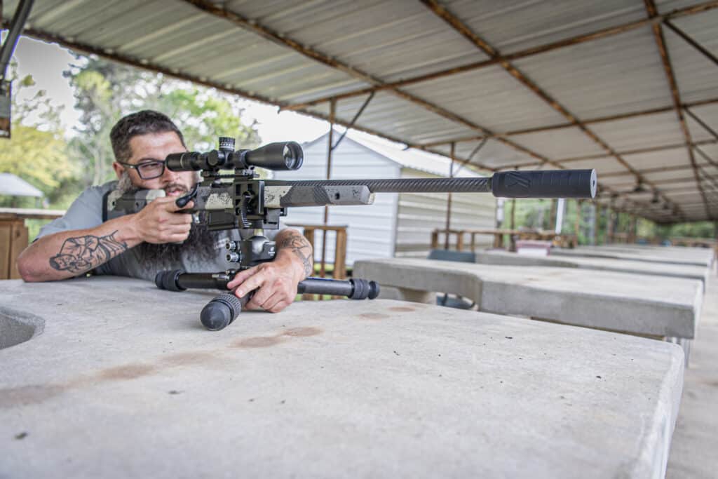 Man wearing glasses aiming a scoped rifle from a prone position at an outdoor covered shooting bench.
