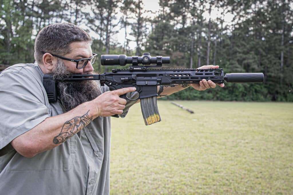 Bearded man wearing glasses aims a scope-equipped black rifle outdoors in a grassy field, with trees in the background.
