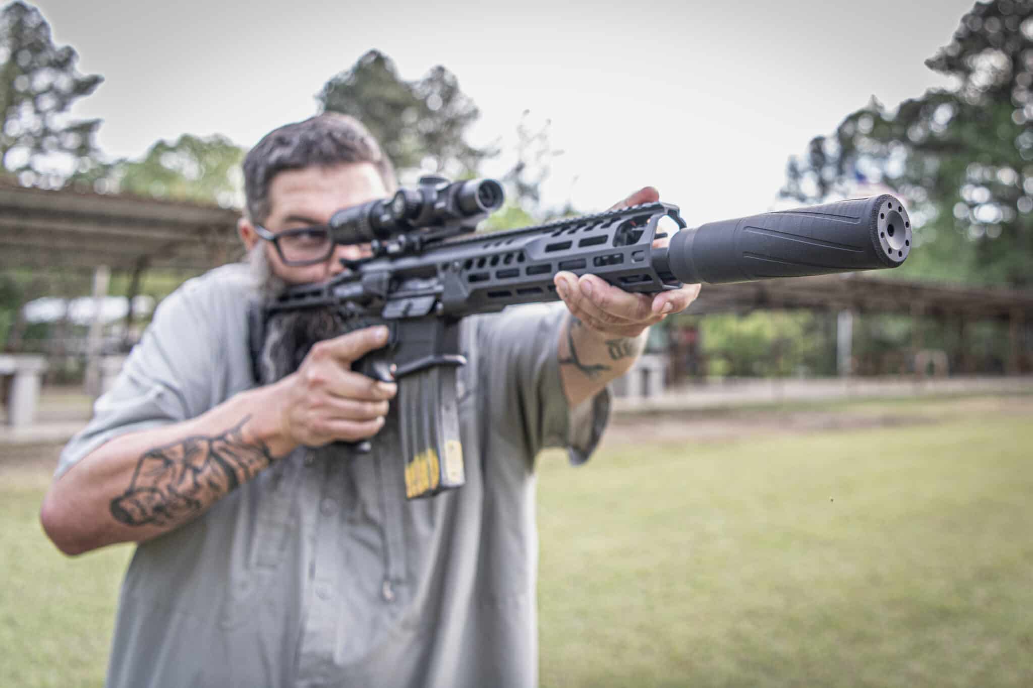 Man aiming a black tactical rifle with a scope, outdoors, with the muzzle to the right and a blurred background.