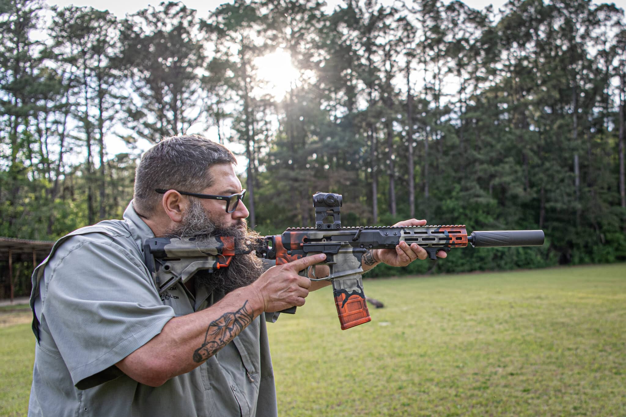 Bearded man wearing sunglasses aiming a camouflaged rifle with an orange grip in a grassy outdoor area, trees in the background.