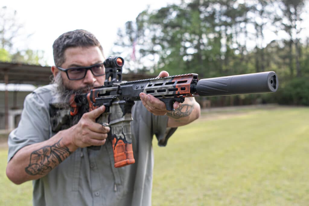 Man with glasses and a beard aims a tactical rifle with a suppressor outdoors on a grassy field.