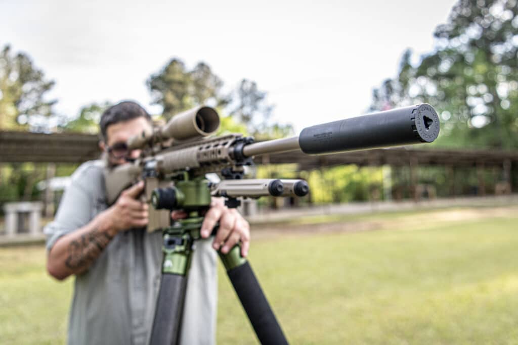 Person aiming a scoped sniper rifle outdoors, focusing on the target.