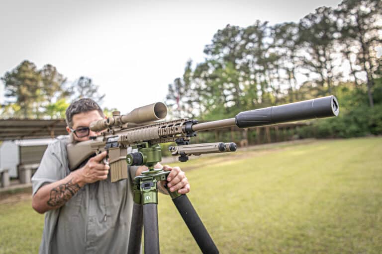 Man wearing glasses aiming a tan precision rifle with a scope outdoors, supported by a bipod on a tripod field setup