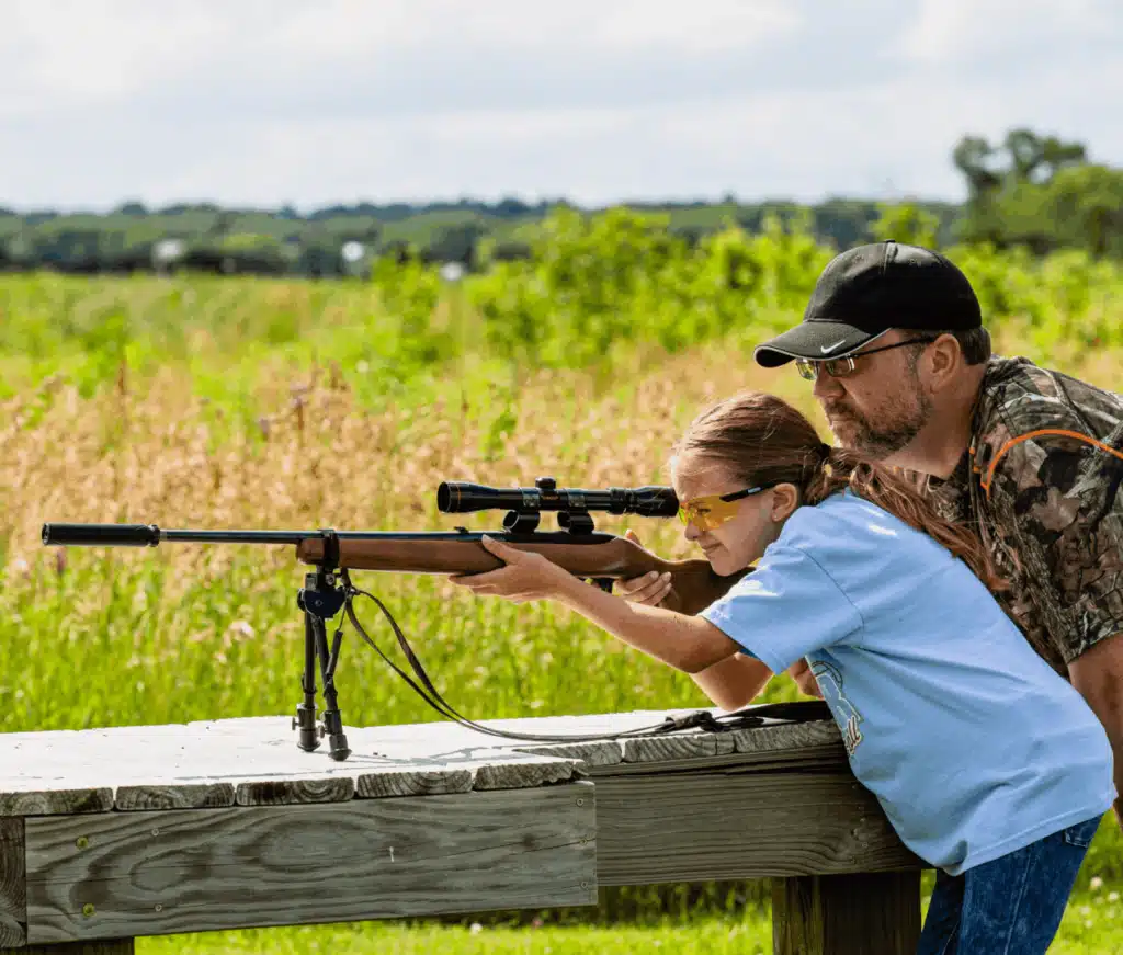 Silencer Central Youth Shooting2 - PopularSuppressors Young girl, wearing safety glasses, aims a scoped rifle on a wooden shooting bench, supervised by a man in camouflage outdoors.