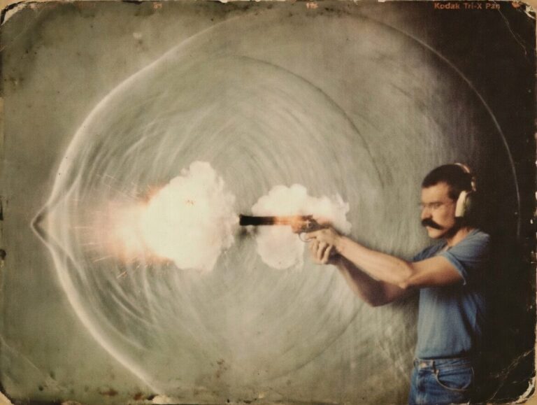 Man with a mustache wearing headphones fires a handgun, muzzle flash and smoke erupting from the barrel to the left.