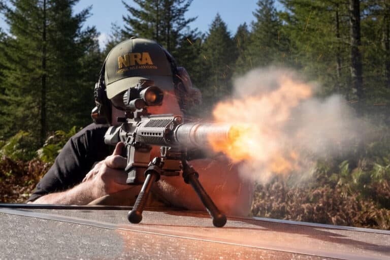 Man in a baseball cap and ear protection fires a rifle from a prone position on a vehicle hood, muzzle flash and smoke visible among trees in a forest setting.