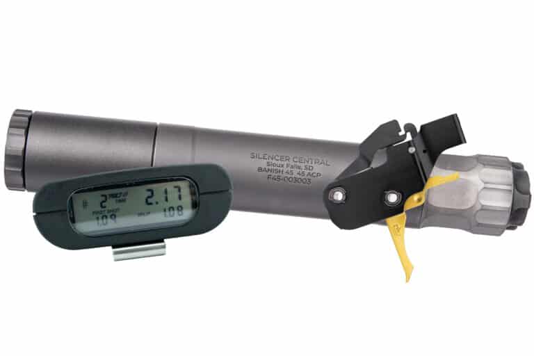 Close-up of a tactical rifle scope with an attached digital display/readout and a yellow trigger-style lever on a grey rifle component. Note: The image shows a firearm accessory with a small electronic timer/display unit attached to the scope.