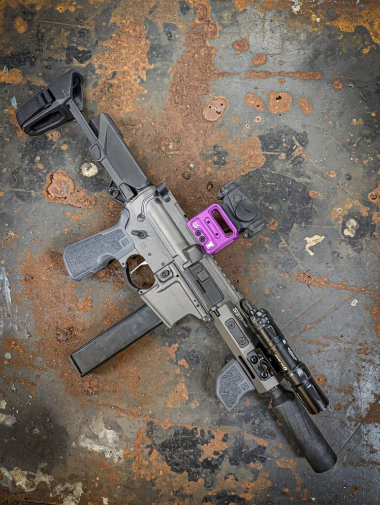 AR-style rifle with a black suppressor, purple red-dot sight, foregrip, and mounted flashlight resting on a rust-stained metal surface.
