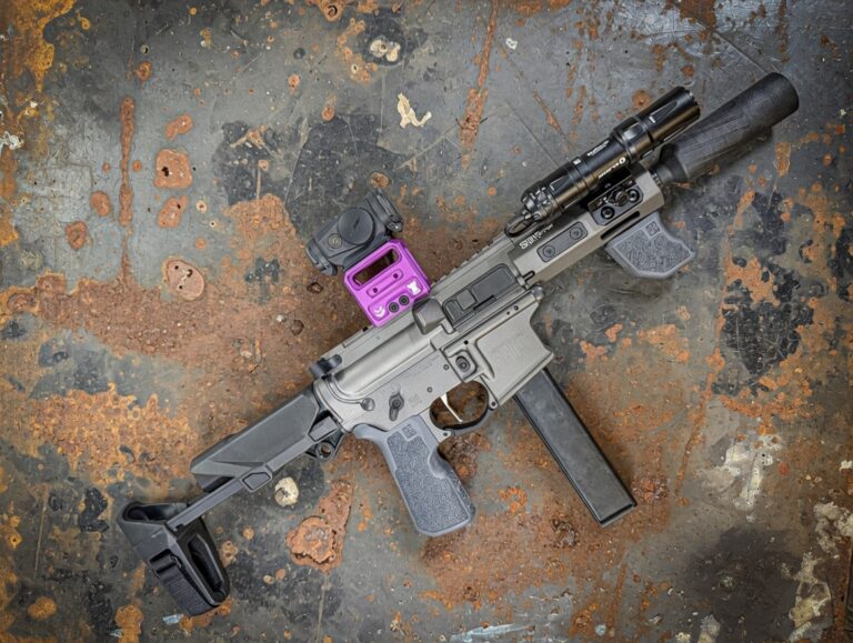 AR-style rifle with a black suppressor, purple red-dot sight, foregrip, and mounted flashlight resting on a rust-stained metal surface.