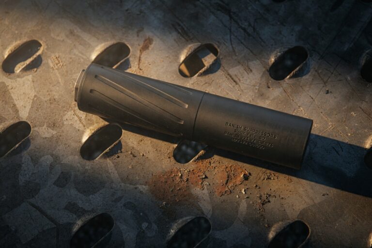 Black firearm suppressor lying on a weathered metal surface with circular holes in view, showing its tapered design.