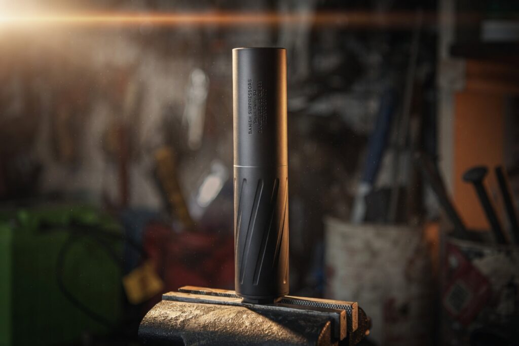 Tall black metal cylinder upright on a rough metal anvil in a cluttered workshop, warm light to the left.