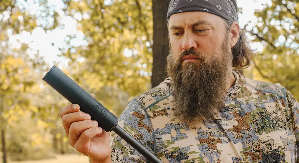BUCK-30-Gallery-1png - PopularSuppressors Bearded man in camouflage garment outdoors, inspecting a black firearm suppressor attached to a barrel.",