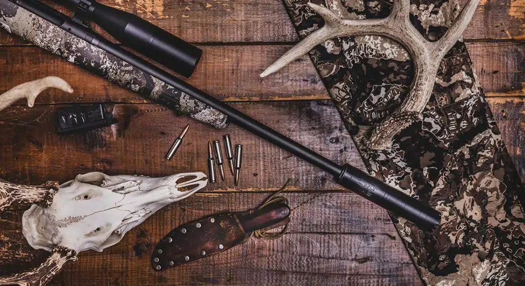 BUCK-30-Gallery-5png - PopularSuppressors Hunting gear laid out on a wooden table: rifle with scope, ammo, deer skull and antlers, camo fabric, and a leather knife sheath.