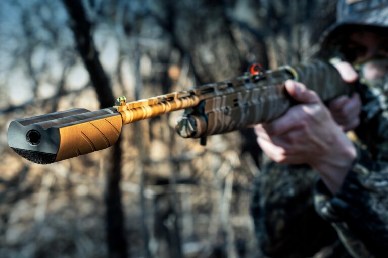 Person in camouflage aiming a tactical rifle with a long barrel in a forest setting.