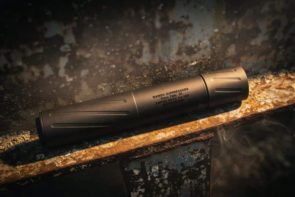 Black cylindrical suppressor resting on a rusted metal ledge, with faint smoke and the label'BANISH SUPPRESSORS' visible on its body