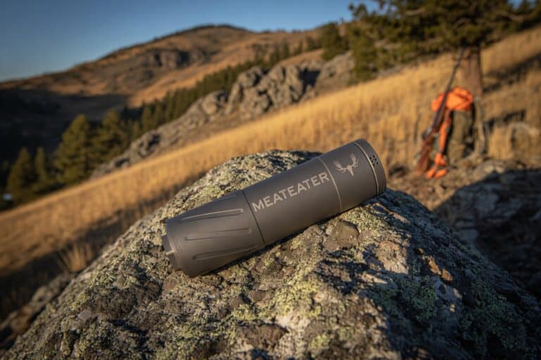 Outdoor scene with a gray Meateater container resting on a textured rock, mountains and backpack in the background.