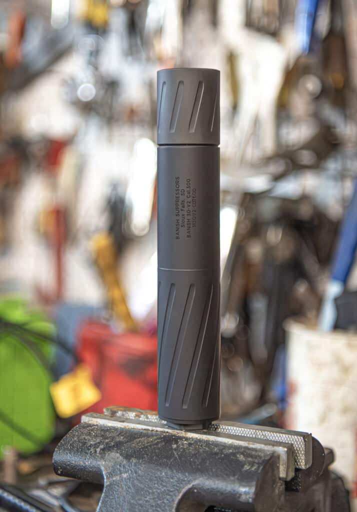 A cylindrical metal tool piece with diagonal grooves stands upright in a bench vise in a cluttered workshop backdrop.