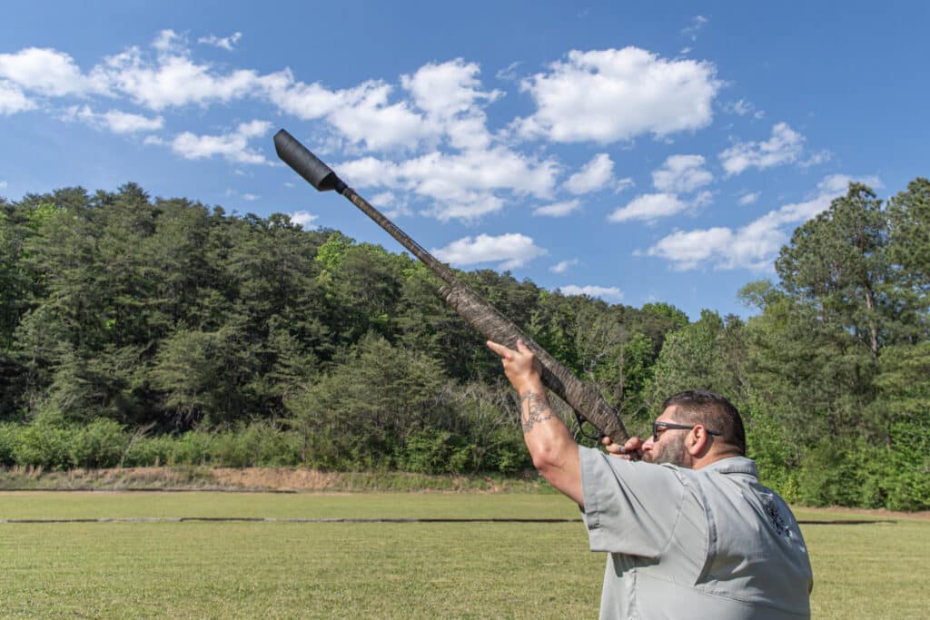 Man with sunglasses fires a shotgun outdoors, raised toward the sky, in a grassy field with trees behind him.