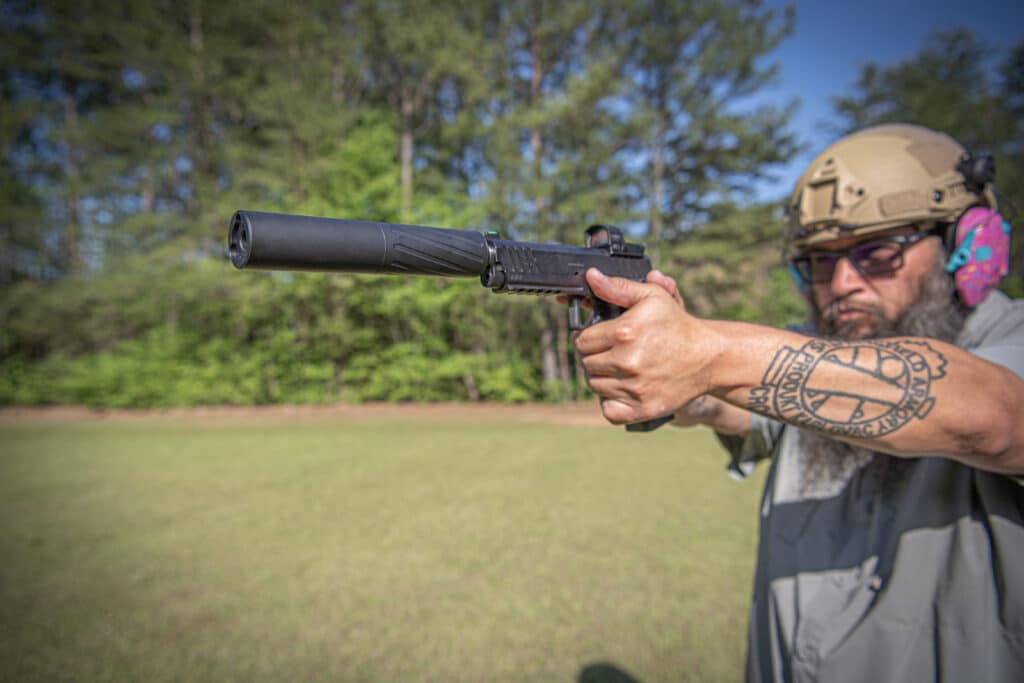 Man in tactical gear and helmet aiming a rifle with a suppressor in a grassy outdoor area.