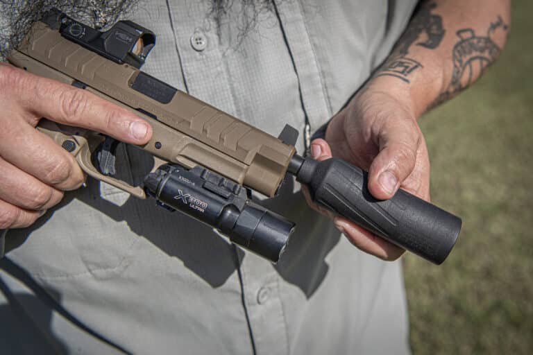 Close-up of a person holding a tan semi-automatic rifle with a red-dot sight and foregrip outdoors, tattoos visible on the arm.