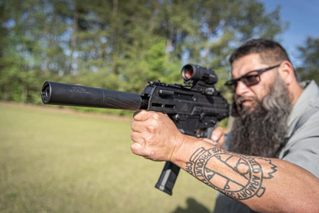 Man with a beard aiming a scoped rifle outdoors, close-up of gun and his tattooed forearm