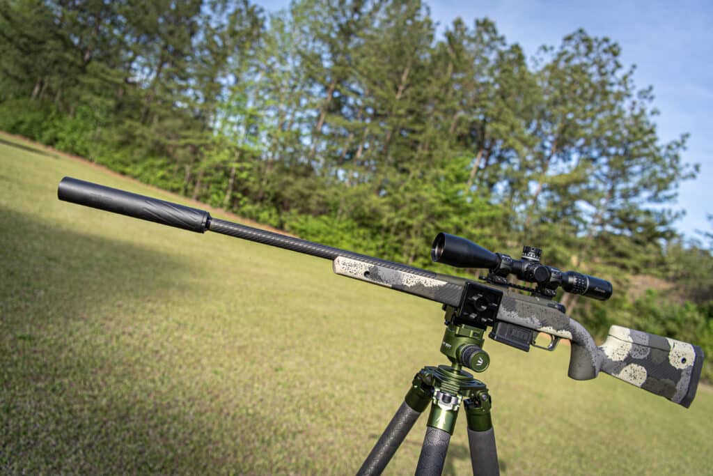 Camouflage-patterned rifle with scope mounted on a bipod in a sunny grassy field with trees in the background.