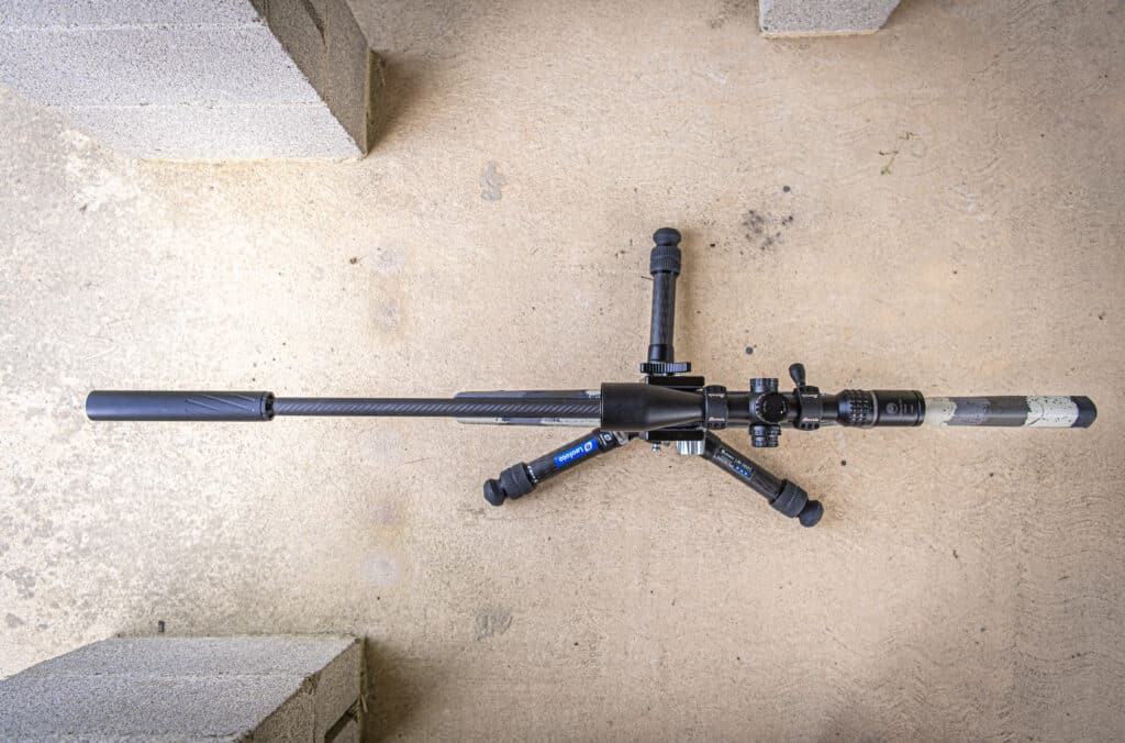 Top-down view of a scoped rifle with a bipod resting on a concrete surface.
