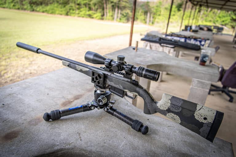 Precision sniper rifle with a scope on a concrete bench at an outdoor shooting range, with targets and greenery in the background.