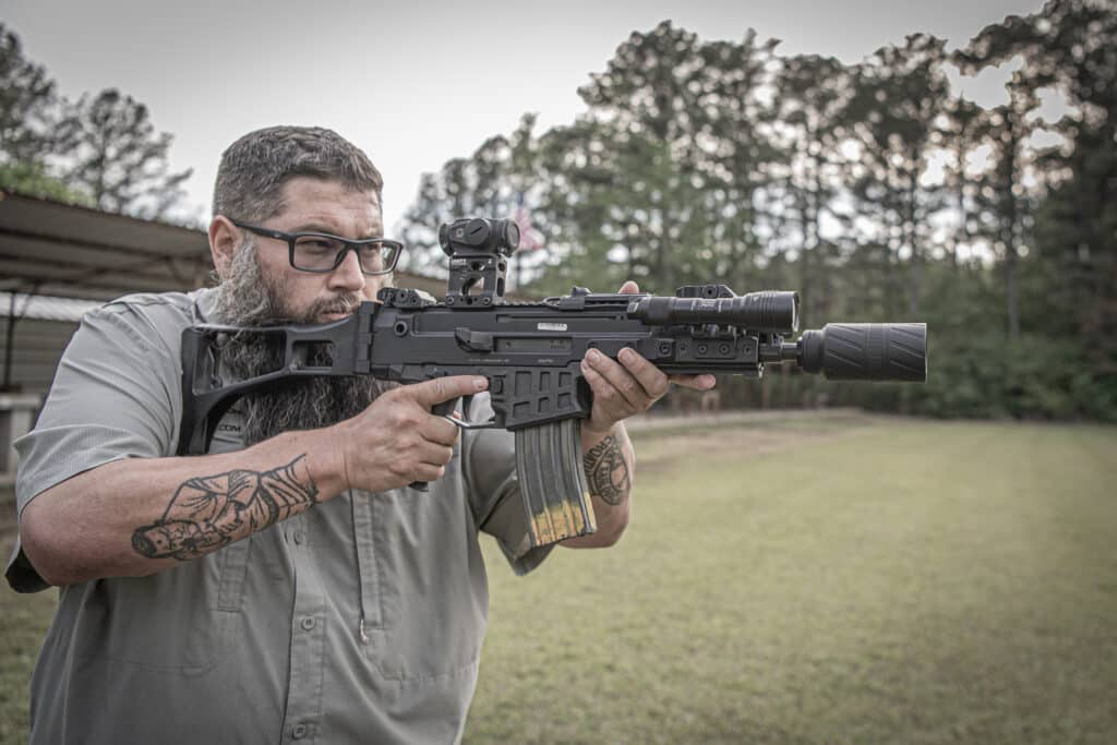 Bearded man in glasses aiming a black tactical rifle with a scope outdoors.