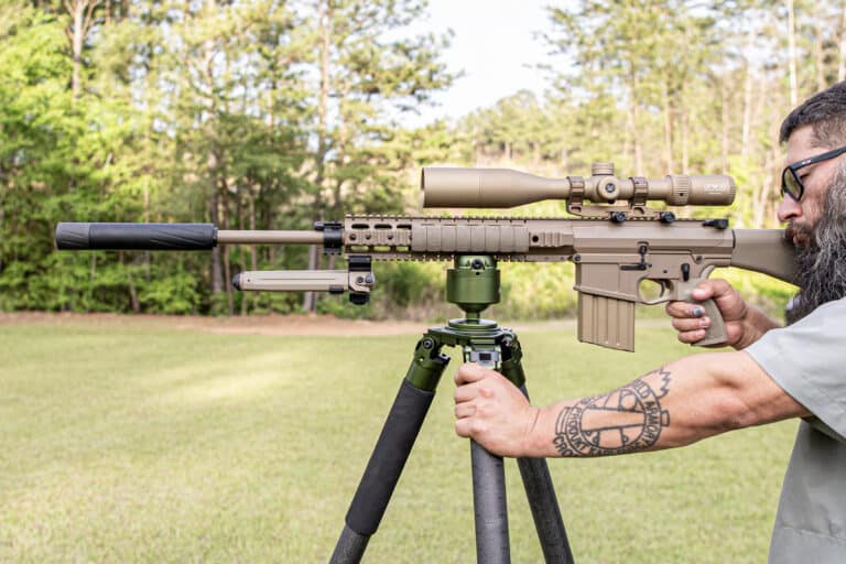 A man with a beard aiming a tan sniper rifle mounted on a bipod in a grassy outdoor setting.