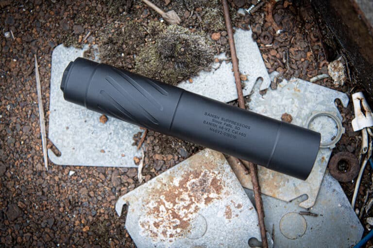 Black rifle suppressor resting on weathered metal plates and rocky ground outdoors, with visible text markings on the cylinder.